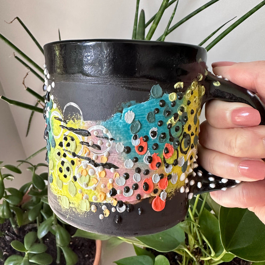 A close up image of a hand holding a black coffee mug with colorful designs and plants in the background